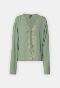 Grønn kortermet bluse i lett stoff, med V-hals, knyteinngang og broderte blomsterdetaljer langs fronten. Lange ermer.
