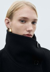 Black high-collared coat made of wool blend, featuring a smooth texture, black buttons, and large gold hoop earrings showcased against a neutral background.
