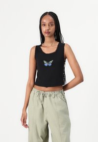 Černý crop top s vroubkovaným výstřihem, na kterém se nachází vyšívaný motýl v modré a zelené barvě uprostřed. Nosený s světle zelenými kalhotami.