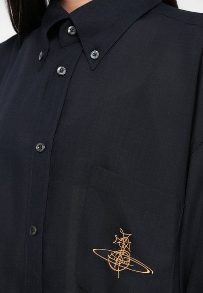 Camisa azul-escura de botão em tecido leve com colarinho pontudo, apresentando um bolso no peito adornado com bordados dourados de um design geométrico.