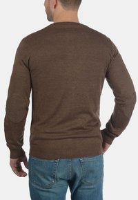 Brun strikkegenser med rund halslinning, ribbede mansjetter og nederkant, samt albuelapper. Stoffet er glatt og noe tettsittende, kombinert med blå jeans.
