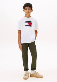 Dreng står med den ene hånd i lommen iført hvid grafisk t-shirt, olivengrønne bukser, hvide sokker og beige sneakers foran en hvid baggrund.