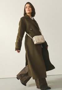 Manteau vert olive à double boutonnage avec tissu texturé ; associé à un pantalon large imprimé léopard marron et un sac bandoulière matelassé crème.