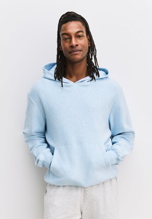 Junger Mann mit langen Dreadlocks, der einen hellblauen Hoodie und graue Jogginghosen trägt, steht mit den Händen in den Taschen vor einer weißen Wand.