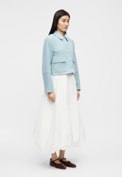 Moteris stovi dėvėdama šviesiai mėlyną trumpą švarkelį, ilgą baltą plazdančią sijoną ir tamsiai rudos spalvos Mary Jane stiliaus batus, ant vientisos fono.