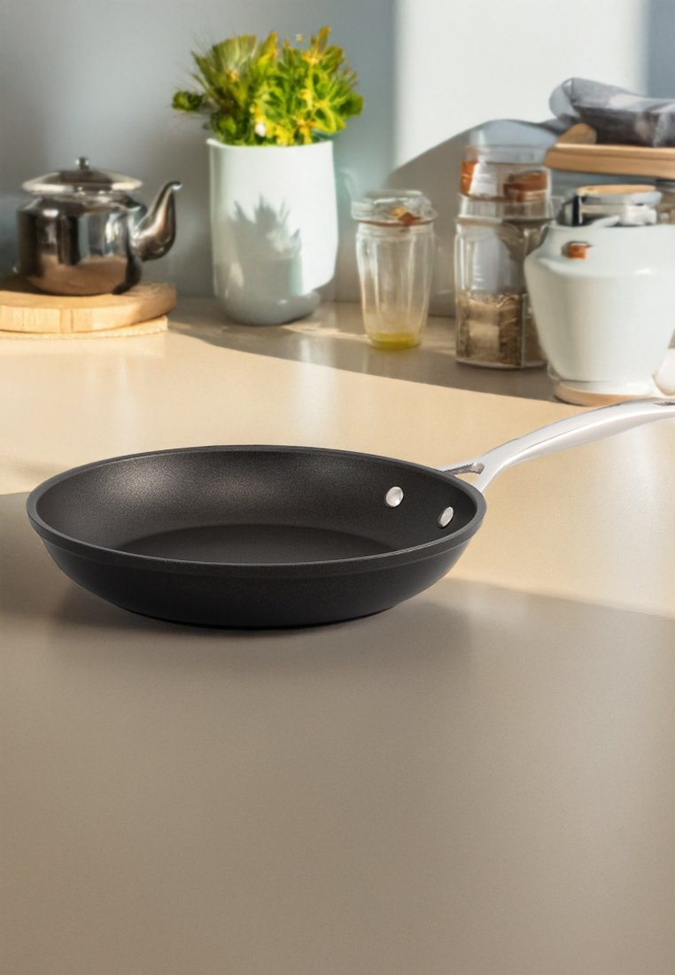 Czarna nieprzywierająca patelnia z gładką powierzchnią, białym uchwytem i dwoma małymi otworami do zawieszenia. Ustawiona na jasnym blacie.