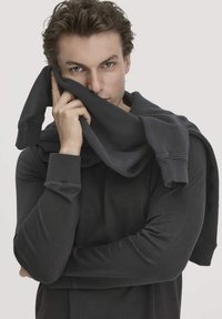 Dunkelgrauer Pullover aus einem weichen, strukturierten Stoff, über die Schultern drapiert. Das Model hält einen Teil des Stoffs an das Gesicht, um das Design und die Passform zu präsentieren.