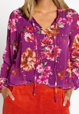 Vrouw met een paarse bloes met roze, rode en oranje bloemen, gecombineerd met feloranje gestructureerde broek, handen in de zakken.