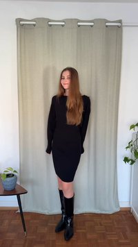 Svart klänning med långa ärmar, figursydd siluett och fyrkantig halsringning. Modellen bär knähöga svarta stövlar. Bakgrunden består av ett ljust draperi och en växt.