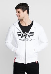 Biela mikina na zips s čiernym logom a pruhmi na prednej strane, predné vrecká a rebrované manžety. Nosená cez čierne tričko.