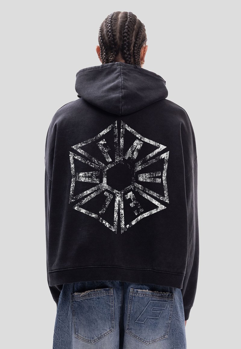 Personne aux cheveux tressés portant un sweat à capuche noir avec un logo géométrique blanc et un texte estompé dans le dos, associé à un jean en denim ample.