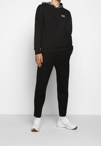 Svart huva med en logotyp, med ett mönstrat huva och uppvikta ärmar. Tillsammans med svarta joggers och vita sneakers.