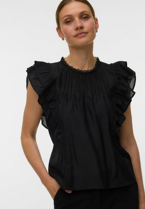 Femme portant un chemisier noir à manches volantées et bordure en dentelle, posant avec une main dans la poche sur un fond uni.