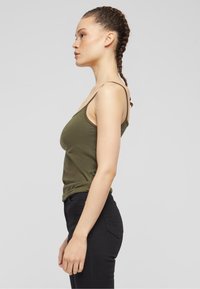 Oliwkowo zielony, dopasowany top na ramiączkach, wykonany z miękkiej bawełny. Połączony z czarnymi dżinsami, pokazany z profilu na jasnym tle.