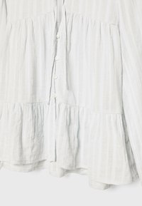 Vit, randig, knapp-up blus gjord av texturerad tyg, med en lagerdesign och en knytning vid halsen.