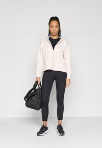 Ροζ φούτερ με φερμουάρ πάνω από μαύρη μπλούζα, μαύρα κολάν, μαύρα αθλητικά παπούτσια και μαύρη τσάντα duffel. Διαθέτει ριγέ υφή και κορδόνια.