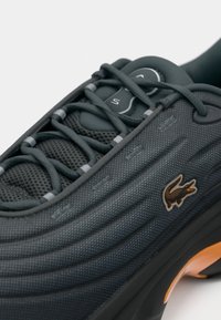 Sorte teksturerede sneakers med tykke snørebånd, mesh tunge, broderet krokodillelogo og orange accent på sålen.