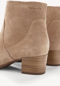 Bottines beige en daim avec talons blocs empilés en bois, coutures visibles et "Tamaris" embossé près du bord supérieur.