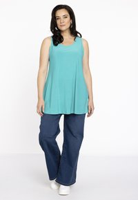 Ein türkisfarbener ärmelloser Top mit lockerem Schnitt, kombiniert mit dunkelblauen weiten Jeans und weißen Sneakers. Der Stoff wirkt weich und glatt.