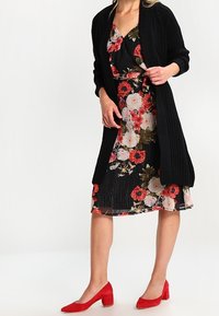 Black knitted cardigan over a floral dress with red and white flowers on a black background. Red heels complete the outfit.