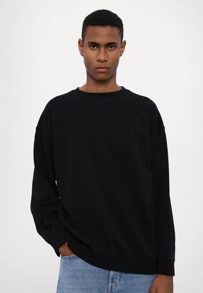 Sweatshirt noir surdimensionné avec un col rond, des épaules tombantes et des poignets côtelés. Porté avec un jean en denim bleu clair. Texture lisse.