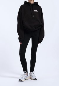 Černá oversized mikina s kapucí a přední kapsou, s bílým logem, kombinovaná s černými legínami a bílými teniskami s šedými akcenty.