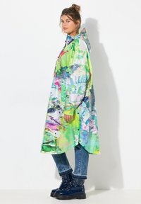 Barevný dlouhý plášť do deště s kapucí, zdobený abstraktním vzorem v zelené, modré a růžové barvě. Stylizovaný přes džíny a černé bojové boty.