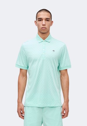 Joven con una camiseta polo verde menta con estampado y un pequeño logo negro en el pecho, de pie frente a un fondo liso.