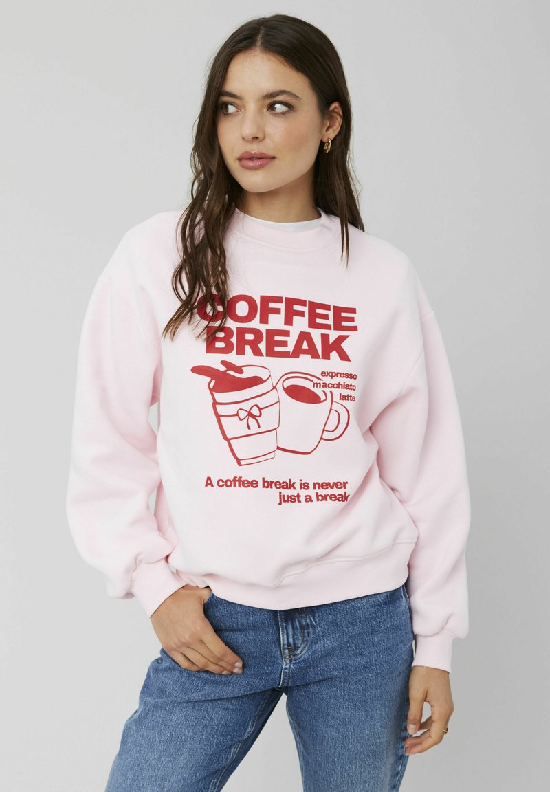 Sweat-shirt rose clair affichant un graphique rouge vif avec "COFFEE BREAK", des images de tasses de café et un slogan en dessous. Coupe décontractée avec des poignets côtelés.
