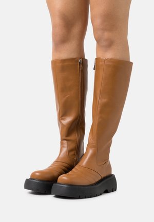 Bottes marron montant jusqu'aux genoux avec une finition en cuir lisse, dotées d'une fermeture éclair latérale, d'accents nervurés sur la pointe et d'une épaisse semelle plateforme noire.