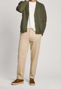Cardigan texturizzato verde oliva, t-shirt bianca, pantaloni beige, sneakers in suede marroni; bottoni sul cardigan, due tasche sui lati.