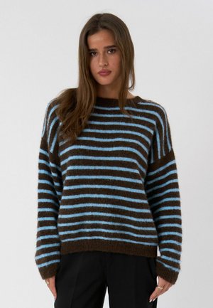 Žena nosi hnedý a svetlomodrý pruhovaný chlpatý sveter s dlhými rukávmi, v kombinácii s čiernymi nohavicami, stojaca pred jednoduchým pozadím.