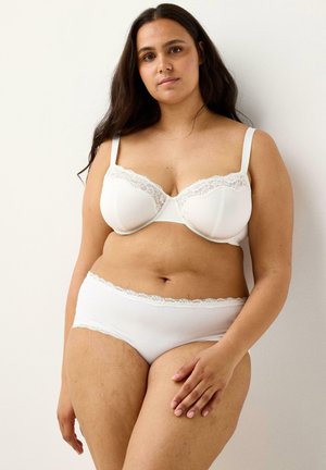 Donna in piedi contro un muro bianco, che indossa un reggiseno bianco con pizzo e mutandine abbinate, con lunghi capelli scuri e un'espressione rilassata.