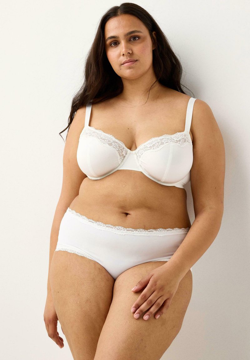 Donna in piedi contro un muro bianco, che indossa un reggiseno bianco con pizzo e mutandine abbinate, con lunghi capelli scuri e un'espressione rilassata.