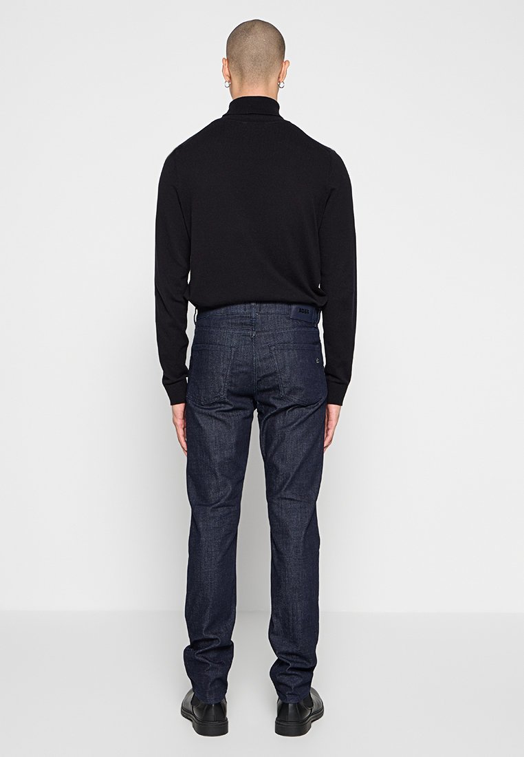 Schwarzer Rollkragenpullover und dunkelblauer Jeans. Das Outfit zeichnet sich durch eine taillierte Passform und glatte Textur aus, mit minimaler Detailgestaltung.