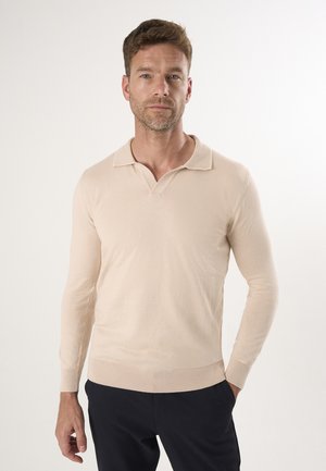 Homme portant un polo beige à manches longues avec un pantalon noir, debout devant un fond clair uni.