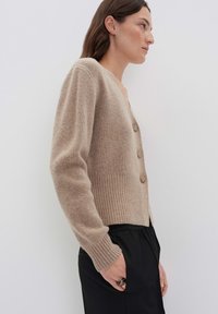 Femme portant un cardigan tricoté beige à boutons avec des poignets et une taille côtelés, associé à un pantalon noir, se tenant de profil avec une main dans la poche.