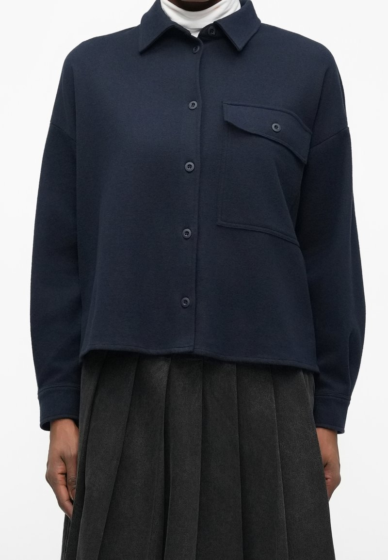Chemise bleu marine boutonnée avec une poche à rabat boutonnée sur la poitrine, portée sur un col roulé blanc et associée à une jupe plissée foncée.