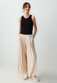 Zwarte geribbelde mouwloze top gecombineerd met beige geplooide wijde broekspijpen. Model draagt witte sneakers. Zachte, vloeiende stoffen met zakken.