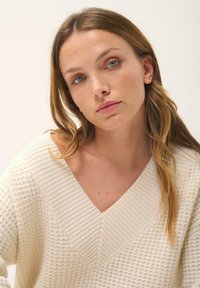 Cremfarbener Strickpullover mit V-Ausschnitt und strukturiertem Muster; weicher, lässiger Schnitt; lange Ärmel; schlichtes Design ohne sichtbare Hardware.