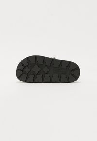 Black footwear sole with deep treads, featuring angular patterns, a smooth edge, and a silver metallic accent near the heel.