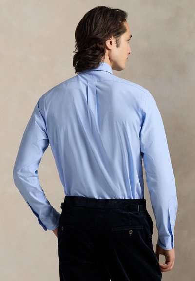 Ανοιχτό γαλάζιο πουκάμισο με μακριά μανίκια, πιέτα στην πλάτη, κλασικό γιακά και μανσέτες με κουμπιά, συνδυασμένο με σκούρο παντελόνι από κορδόνι.