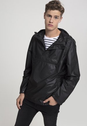 Veste à capuche noire en matériau imperméable, dotée d'une poche zippée sur le devant, de poignets élastiques et portée par-dessus une chemise rayée.