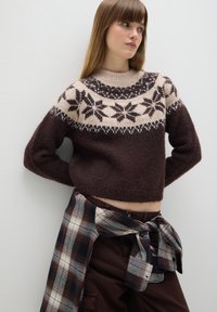 Bruine gebreide trui met een patroon in roomkleur en donkerbruin op de schouders. Gekleed met een geruite blouse om de taille en donkere broek.