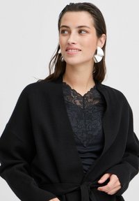 Melnā apmalejuma kardigāns, kas izgatavots no mīksta materiāla, ar sasienamu aizdari. Zem tā ir pieguļošs melns tops ar mežģīņu apkakles detaļu. Metāliskas auskari.