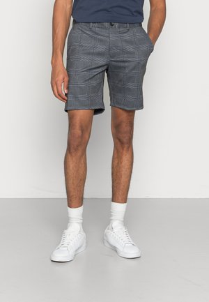 Homme portant un short slim gris à carreaux, des chaussettes blanches, des baskets blanches et une chemise bleu foncé, debout avec les mains dans les poches.
