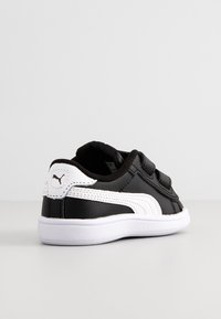 Baskets Puma noir et blanc avec semelle blanche, munies de deux bandes velcro et du logo Puma sur la languette du talon, vues de l'arrière côté gauche.