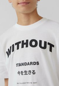 Witte katoenen T-shirt met vetgedrukte zwarte tekst "WITHOUT STANDARDS" en extra Japanse tekst. Ronde halslijn en een losse pasvorm.