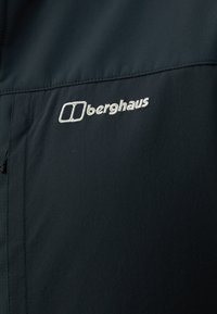 Musta takki, jossa on valkoisella brodeerattu "Berghaus"-logo. Materiaali on sileää hienovaraisella tekstuurilla ja siinä on vetoketjullinen tasku.