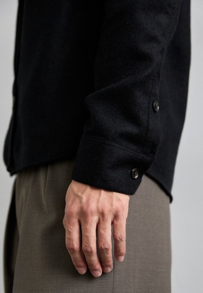 Black wool shirt with a textured finish and buttoned cuffs, worn over beige trousers. Features a tailored fit and clean design.